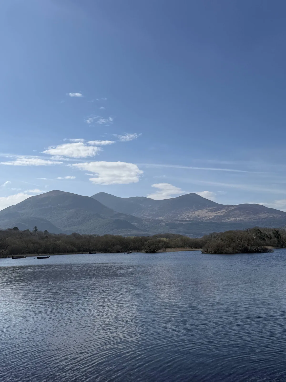 Blue lake water with small boats in front of Irish mountains under a clear spring sky.